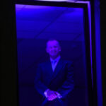 Na obrazie widać sylwetkę mężczyzny w garniturze, oświetlonego fioletowym światłem. Tło jest ciemne. Postać stoi w prostokątnym oknie lub ramach.