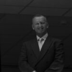 Mężczyzna w garniturze i krawacie, uśmiecha się, stojąc na ciemnym tle z geometrycznym wzorem na suficie.