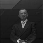 Czarno-białe zdjęcie przedstawia mężczyznę w eleganckim garniturze na tle sufitu z kwadratowymi panelami. Mężczyzna stoi z rękami złożonymi na wysokości pasa. Oświetlenie jest miękkie, a atmosfera formalna.
