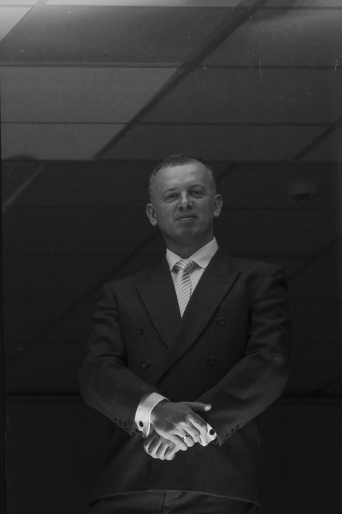 Czarno-białe zdjęcie przedstawia mężczyznę w eleganckim garniturze na tle sufitu z kwadratowymi panelami. Mężczyzna stoi z rękami złożonymi na wysokości pasa. Oświetlenie jest miękkie, a atmosfera formalna.