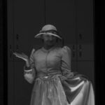 Osoba w kapeluszu i długiej sukni pozuje z uniesioną ręką. Zdjęcie jest czarno-białe. W tle widać szafki.