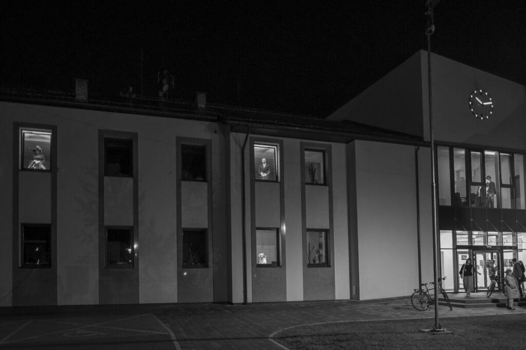 Budynek nocą z jasno oświetlonymi oknami. W oknach widać sylwetki ludzi. Na ciemnym tle widać zegar i rowery.