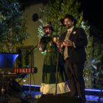 Dwie osoby śpiewają na scenie wieczorem, w tle zielone drzewka. Jedna w zielonej sukni, druga w garniturze i kapeluszu. Obok klawisze z napisem Blues Junkers.