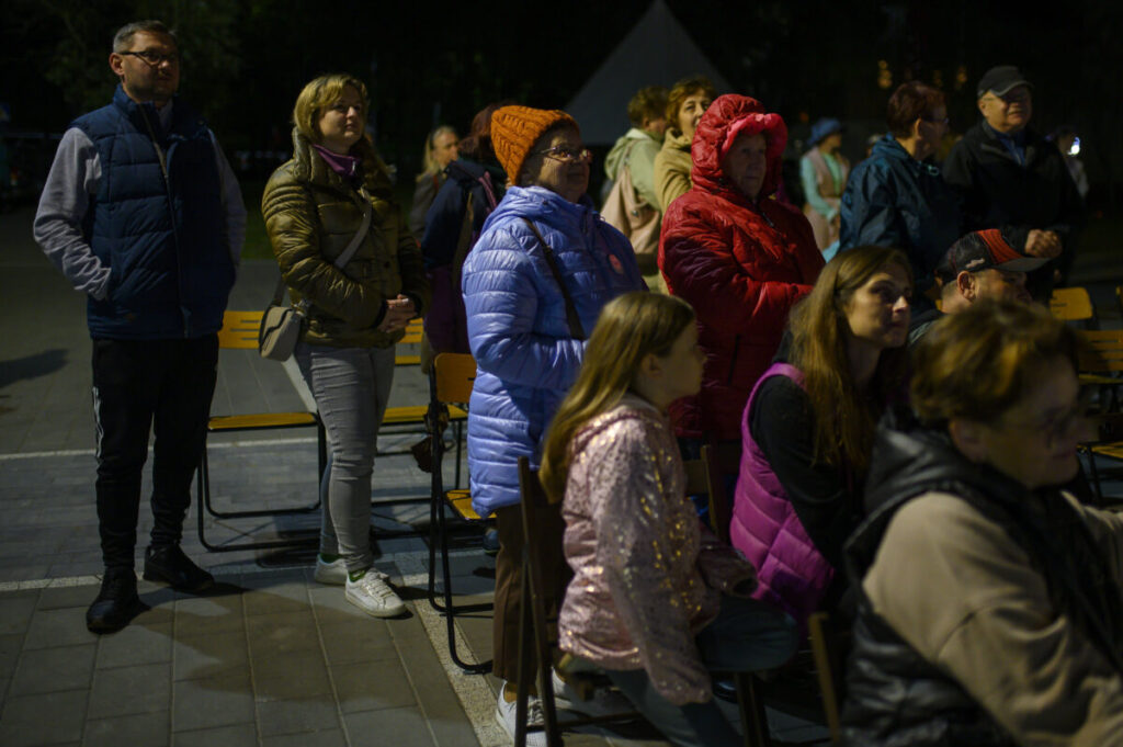 Grupa ludzi w zimowych kurtkach stoi na zewnątrz. Kobiety i mężczyźni w kolorowych ubraniach, na tle ciemnego nieba. Sceneria nocna, obok chodnik.