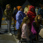 Grupa ludzi w zimowych kurtkach stoi na zewnątrz. Kobiety i mężczyźni w kolorowych ubraniach, na tle ciemnego nieba. Sceneria nocna, obok chodnik.