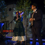 Dwoje ludzi na scenie nocą, ubranych elegancko; kobieta trzyma miskę. Tło to drzewa z kolorowym oświetleniem.