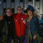 Trzy osoby pozują wieczorem: kobieta w kapeluszu, mężczyzna w czerwonej kurtce, kobieta w czarnej kamizelce. Tło szaro-niebieskie z widocznym starym autem. Kobieta trzyma parasolkę, klimat retro.