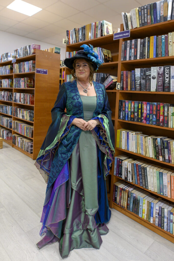 Osoba w eleganckiej sukni z kapeluszem znajduje się w bibliotece. Suknia w zieleni i fioletach harmonizuje z książkami na półkach. Tło stanowią regały z książkami oznaczonymi kategoriami.