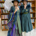 Dwie kobiety w zielonych sukniach retro pozują w bibliotece. Jedna trzyma koronkowy parasol. W tle półki z książkami.