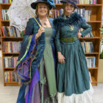 Dwie kobiety w eleganckich, kolorowych sukniach stoją przed półką z książkami. Pierwsza trzyma białą parasolkę, a obie mają kapelusze. W tle regał z literaturą polską.