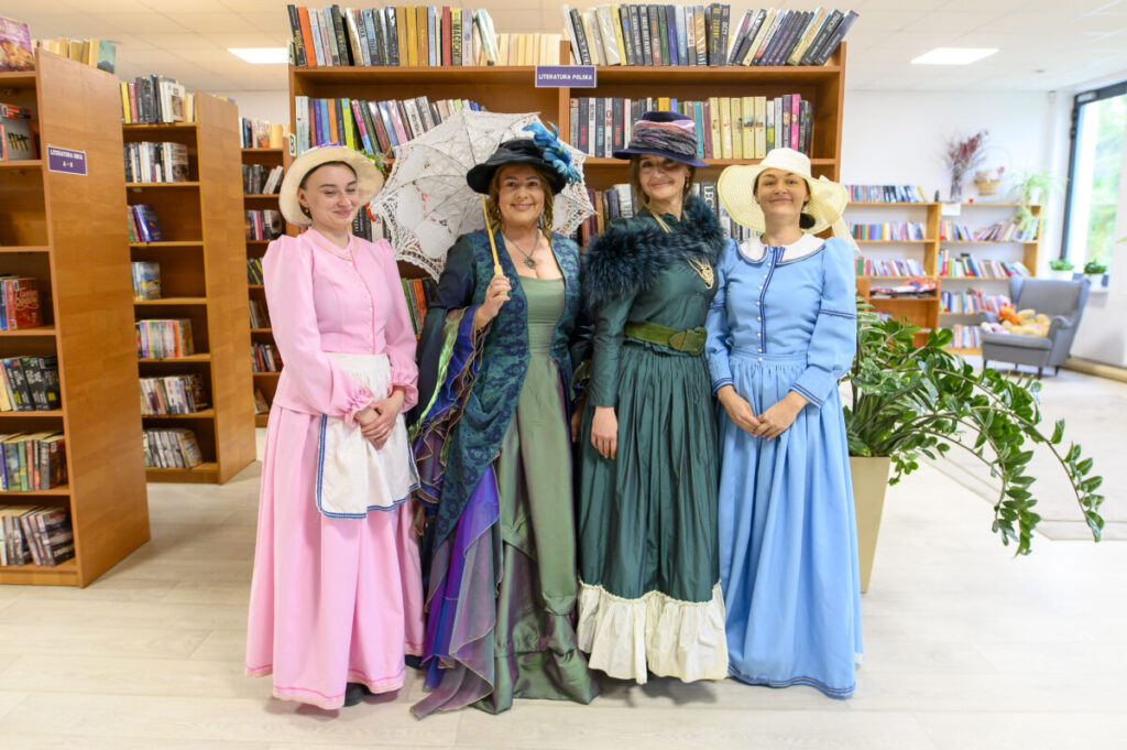 Cztery kobiety w strojach retro stoją w bibliotece. Mają różowe, zielone i niebieskie suknie oraz kapelusze. W tle widoczne są półki z książkami.