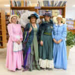Cztery kobiety w strojach retro stoją w bibliotece. Mają różowe, zielone i niebieskie suknie oraz kapelusze. W tle widoczne są półki z książkami.