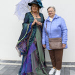 Kobieta w kolorowej sukni i kapeluszu z parasolką, obok starsza pani w niebieskiej kurtce. Stoją na szarej kostce. Tło białe.