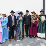 Grupa ludzi w strojach retro pozuje na tle białej ściany. Kolory dominujące to różowy, niebieski i zielony. Kilka osób ma kapelusze.