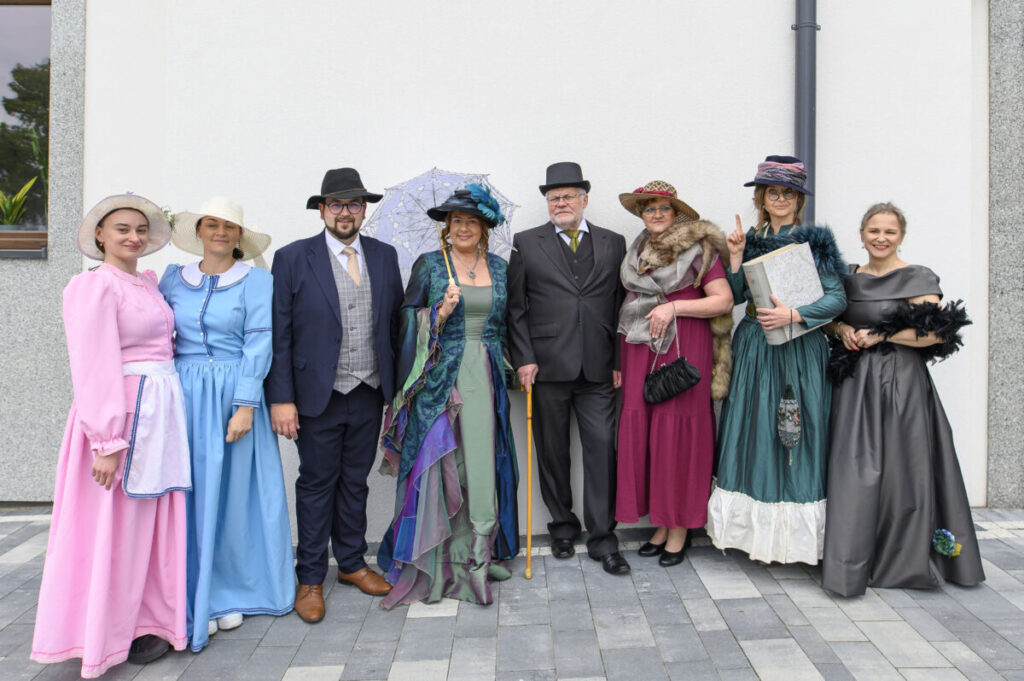 Grupa ludzi w strojach retro pozująca na tle białej ściany. Stroje w kolorach różowym, niebieskim i zielonym. Dwie osoby trzymają parasolkę i książkę.