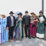 Grupa ludzi w strojach retro pozująca na tle białej ściany. Stroje w kolorach różowym, niebieskim i zielonym. Dwie osoby trzymają parasolkę i książkę.