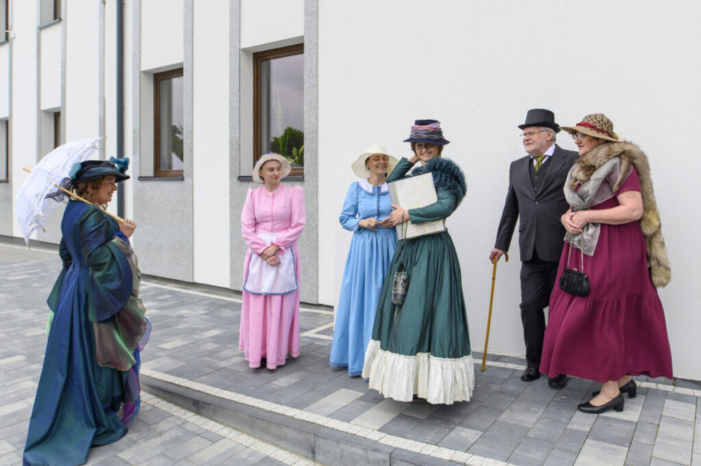 Grupa osób w strojach retro pozuje na tle białej ściany. Kolorowe suknie i kapelusze przyciągają wzrok. Kafelkowana, szara posadzka.