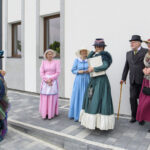 Grupa osób w strojach retro pozuje na tle białej ściany. Kolorowe suknie i kapelusze przyciągają wzrok. Kafelkowana, szara posadzka.