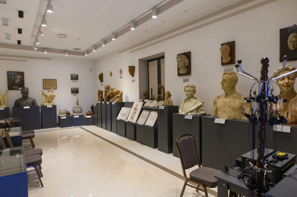 Muzeum z rzeźbami na ciemnych postumentach. Ściany białe, podłoga jasna, oświetlenie punktowe. W tle kilka obrazów i rzeźb.