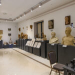 Muzeum z rzeźbami na ciemnych postumentach. Ściany białe, podłoga jasna, oświetlenie punktowe. W tle kilka obrazów i rzeźb.