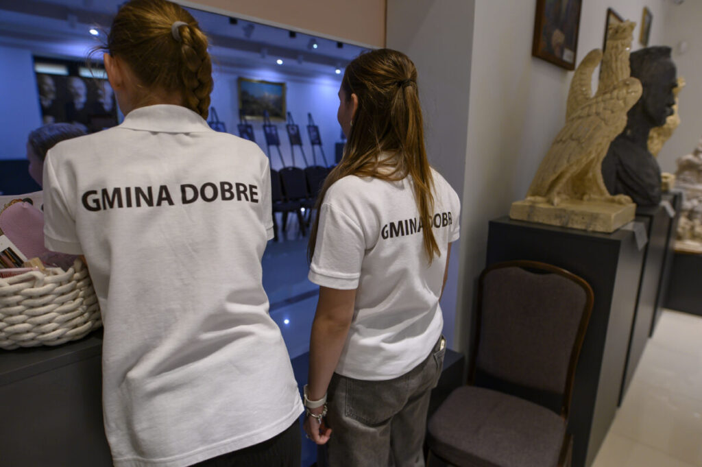 Dwie osoby w białych koszulkach GMINA DOBRE stoją w pomieszczeniu galeryjnym. Obok znajduje się rzeźba, a w tle widoczne są obrazy. Atmosfera jest elegancka i artystyczna.