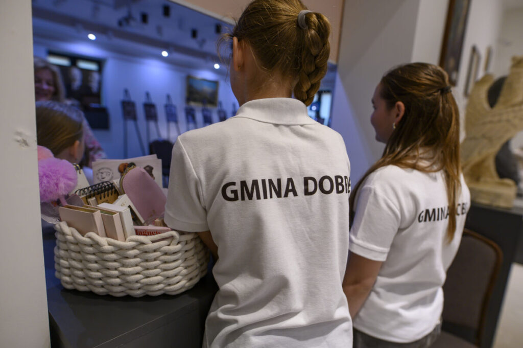 Dwójka dzieci w białych koszulkach, z napisami Gmina Dobre. Na blacie kosz z przedmiotami, w tle galeria.