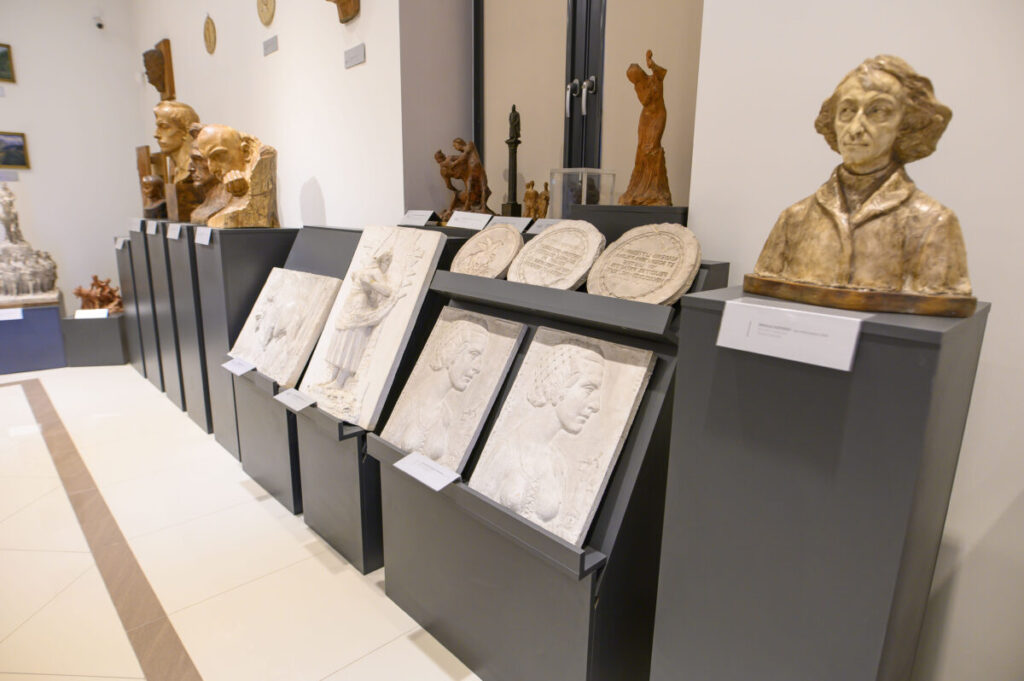 Galeria z popiersiami, płaskorzeźbami i rzeźbami na czarnych postumentach. Jasne ściany, kontrastujące ciemne podstawy. Rzeźby w ciepłych odcieniach brązu i bieli.