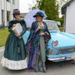 Dwie kobiety w stylowych, historycznych strojach stoją przy błękitnym samochodzie. Jedna trzyma książkę, druga parasolkę. W tle budynek i rowery.
