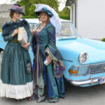 Dwie kobiety w historycznych, kolorowych sukniach stoją obok błękitnego klasycznego auta. Jedna trzyma książkę, druga parasolkę. W tle budynek i trawnik.