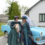 Dwie kobiety w sukniach retro i kapeluszach stoją przed niebieskim, klasycznym samochodem. Jedna trzyma parasolkę, a druga książkę. Tło to jasny dom i drzewa.