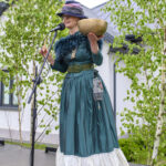 Kobieta w zielonej sukni i kapeluszu stoi na scenie, trzymając złotą misę. Mikrofon przed nią. W tle drzewa i budynek.