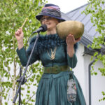 Osoba w zielonej sukni i kapeluszu trzyma misę i pałeczkę. Obok, mikrofon i zielone drzewa. Tło to biały budynek z szarym dachem.