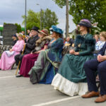 Grupa osób w strojach historycznych siedzi na zewnątrz. Widoczne są kolory różowy, zielony, granatowy. W tle drzewa i chmurne niebo.