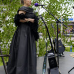 Kobieta w czarnej sukni śpiewa do fioletowego mikrofonu. W tle zielone liście drzew. Obok stoją czarne gitary.