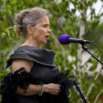 Na zdjęciu kobieta w czarnej sukni i z mikrodzieniem o fioletowej główce śpiewa na zewnątrz. Tło to zielone liście drzewa. Jej bransoletka i kolczyki są zielone.