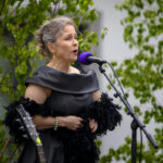 Osoba w czarnej sukience śpiewa do mikrofonu na zewnątrz. Tło zielone, z roślinami. Fioletowy mikrofon dodaje kolor.