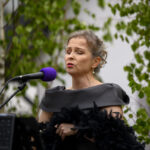 Kobieta w czarnej sukni śpiewa do mikrofonu z fioletową osłoną. W tle zielone liście na jasnym tle. Elegancka i skupiona ekspresja.