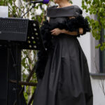 Kobieta w czarnej sukni śpiewa przy mikrofonie na tle zielonych roślin. Ma krótkie, zaczesane włosy. Obok stoi statyw z nutami.