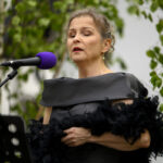 Kobieta śpiewa do fioletowego mikrofonu na tle zieleni. Ubrana w elegancką czarną suknię, trzyma czarny szal. Tło rozmyte z liśćmi.