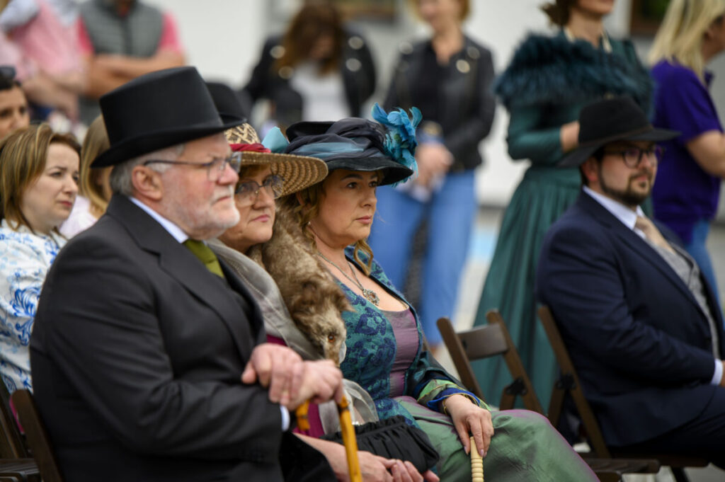 Zdjęcie przedstawia ludzi w eleganckich strojach vintage z kapeluszami, siedzących na krzesłach. Tło jest rozmyte, widać zarys innych osób. Dominują kolory ciemne i zielone.