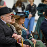 Zdjęcie przedstawia ludzi w eleganckich strojach vintage z kapeluszami, siedzących na krzesłach. Tło jest rozmyte, widać zarys innych osób. Dominują kolory ciemne i zielone.