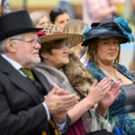 Trzy osoby w eleganckich strojach i kapeluszach siedzą i klaszczą. Mężczyzna w czarnym fraku, kobiety w kolorowych sukienkach. Tło rozmyte, inne osoby słabo widoczne.