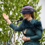 Kobieta w zielonej sukni i kapeluszu śpiewa do mikrofonu. Jej kapelusz ozdobiony jest szalem w pastelowych kolorach. W tle zielone liście na białym tle.