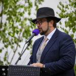 Mężczyzna w kapeluszu przemawia do mikrofonu z fioletową osłoną. Ma na sobie granatowy garnitur. W tle widoczne są zielone liście.