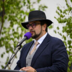 Mężczyzna w kapeluszu przemawia do mikrofonu. Ma na sobie granatowy garnitur i jasny krawat. Tło z zielonymi liśćmi.