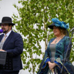 Mężczyzna i kobieta w kapeluszach stoją przy mikrofonach na tle liściastych drzew. Mężczyzna ma ciemny garnitur, kobieta zielony strój. Tło jest jasne.