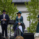 Na zdjęciu widać troje ludzi w strojach retro na tle zielonych drzew. Dwie kobiety w sukniach, a mężczyzna w garniturze przy mikrofonie. W tle biały budynek.