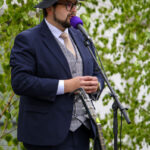 Mężczyzna w garniturze i kapeluszu mówi do mikrofonu. Ma fioletowy mikrofon, w tle zielone liście. Obok stoi gitara.