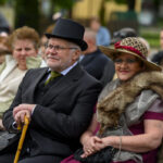 Para w eleganckich, retro strojach siedzi na zewnątrz. Mężczyzna ma czarny frak i cylinder. Kobieta w kapeluszu i futrze, z uśmiechem.