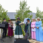 Grupa osób w strojach retro stoi na scenie na tle zielonych drzew. Ubrania są w kolorach niebieskim, różowym i zielonym. W tle białe budynki z szarymi dachami.